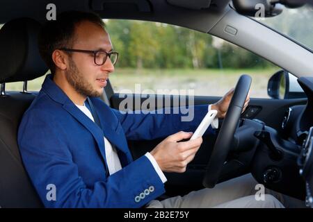 Side view of smiling young business man in glasses sitting in car and typing / messaging on smartphone, stopped so as not to be distracted. Stock Photo