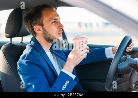 Businessman in a blue blazer holding steering wheel and drinking coffee from a paper cup while driving a car so as not to fall asleep at the wheel, lo Stock Photo