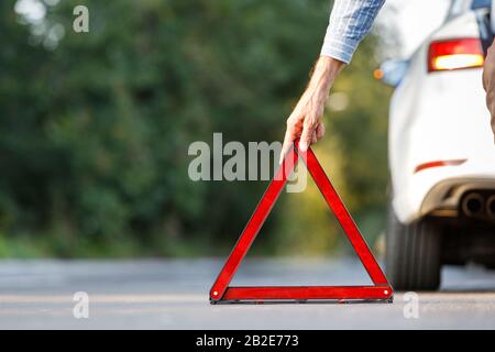 Close up of man driver putting red warning triangle/emergency stop sign behind his broken car on the side of the road, copy space and blurred backgrou Stock Photo