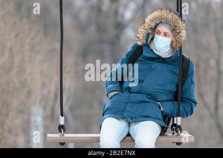 Sick man with a hood sitting alone on a swing, wearing protective facial mask against transmissible infectious diseases and as protection against the Stock Photo