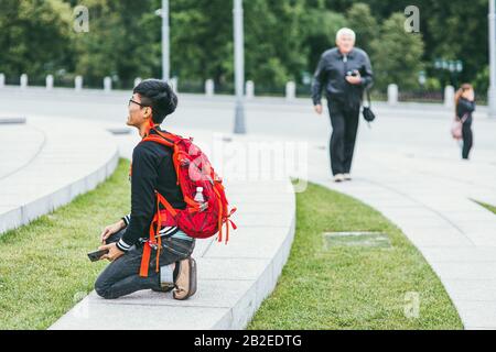 Moscow, Russia - july 7, 2017. A young happy Asian male photographer with a bright backpack on his shoulders photographs the monument. side view Stock Photo