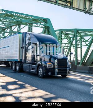 Big rig black stylish diesel long haul industrial semi truck transporting frozen commercial cargo in refrigerator semi trailer running on the arched t Stock Photo