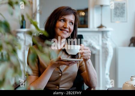 Middle Aged Brunette Woman Drinking Tea From Porcelain Cup And Looking Through Window. Natural Ageing Stock Photo