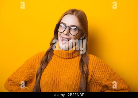 Caucasian girl with red hair and freckles is looking through her glasses while posing on yellow background in a sweater Stock Photo