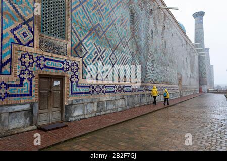 Mother and son in yellow rain jackets in front of colorful wall, snow falling at The Registan, the heart of the ancient city of Samarkand - Uzbekistan Stock Photo