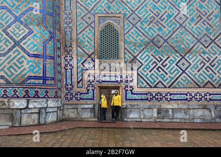 Mother and son in yellow rain jackets in front of colorful wall, snow falling at The Registan, the heart of the ancient city of Samarkand - Uzbekistan Stock Photo
