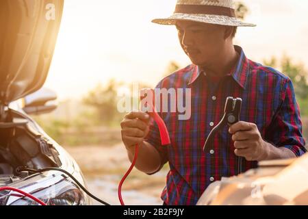 Asian man holding black and red battery cable and trying to connect the cable to his car. Car maintenance before driving concept Stock Photo