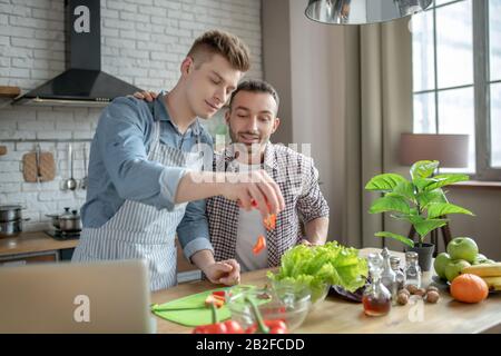 Blond-haired man pouring chopped pepper into a plate, bearded standing nearby Stock Photo