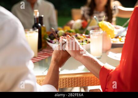 Rear view mid section of a Caucasian couple sitting outside with their family at a dinner table set for a meal, holding hands together in prayer. Fami Stock Photo