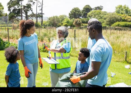 Front view of a senior African American woman standing in a field wearing a hi-vis vest and gloves, holding a clipboard and instructing a diverse grou Stock Photo