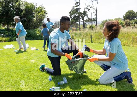 Side view of a young African American man and woman kneeling in a field wearing t shirts with volunteer written on them, holding a rubbish sack and co Stock Photo