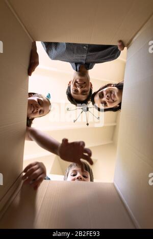 Low angle front view close up of a Caucasian couple and their young son and daughter looking down into an open cardboard box while unpacking at their Stock Photo