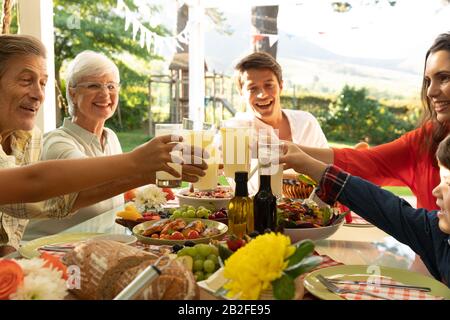 Front view close up of a multi-generation Caucasian family sitting outside at a dinner table set for a meal, smiling, raising glasses together and mak Stock Photo