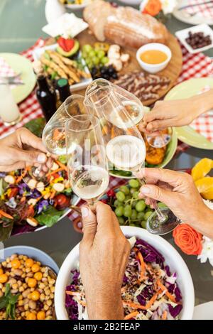 High angle view of the hands of a senior and an adult Caucasian couple holding champagne glasses and making a toast over a table of food outdoors. Fam Stock Photo