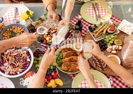 High angle view of the hands of a multi-generation Caucasian family holding glasses and making a toast over a table of food outdoors. Family enjoying Stock Photo