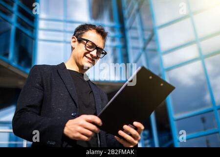 Portrait of successful young handsome engineer, architect, builder, businessman, wearing in a suit holding clipboard, skyscraper background. Stock Photo