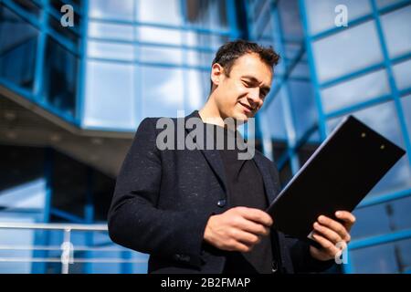 Portrait of successful young handsome engineer, architect, builder, businessman, wearing in a suit holding clipboard, skyscraper background. Stock Photo