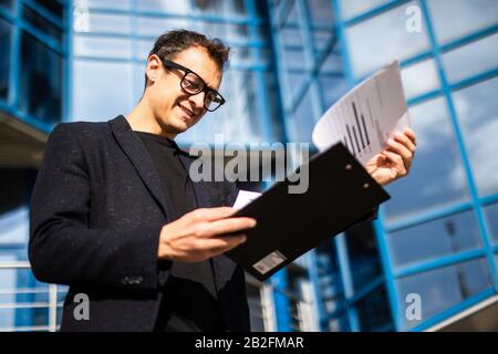 Portrait of successful young handsome engineer, architect, builder, businessman, wearing in a suit holding clipboard, skyscraper background. Stock Photo