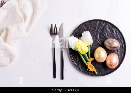 Easter table setting with spring tulips. Stone empty plate, cutlery, napkin and golden eggs on stone Stock Photo