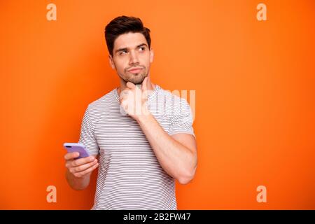 Photo of minded serious millennial guy hold telephone deep think new instagram blog post text arm on chin look side wear striped t-shirt isolated Stock Photo