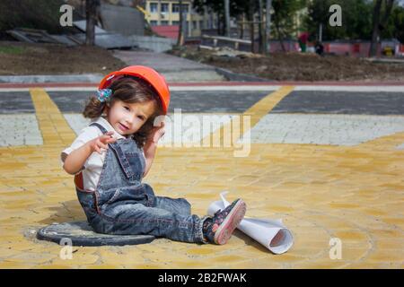 Little baby girl builder with the construction helmet and poster in denim uniform smiling on construction site Stock Photo