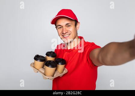 Happy man from delivery service in red t-shirt and cap giving food order and holding coffee boxes over white background Stock Photo
