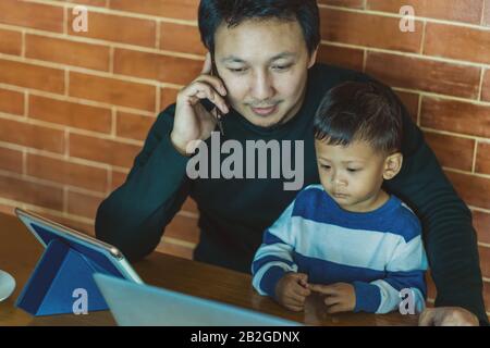 Asian single dad with son are looking the cartoon via technology laptop together with phone calling for work when living in loft house for Self learni Stock Photo