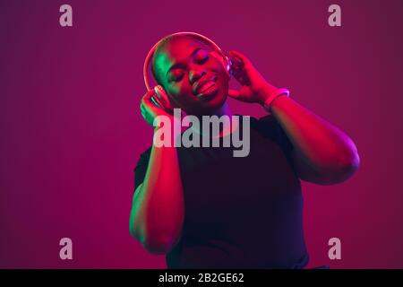 Listen to music. African-american young woman's portrait on purple background. Beautiful model in wireless headphones. Concept of emotions, facial expression, sales, ad, inclusion. Copyspace. Stock Photo