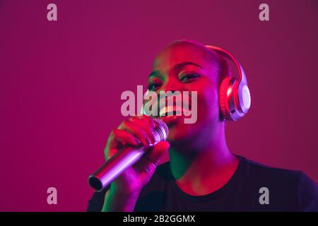 Singing in headphones. African-american young woman's portrait on purple background. Beautiful model in black shirt. Concept of emotions, facial expression, sales, ad, inclusion, diversity. Copyspace. Stock Photo