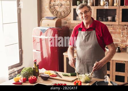 Smiling Aged Man Preparing Fresh Vegetable Salad, Enjoying Cooking In Kitchen Stock Photo