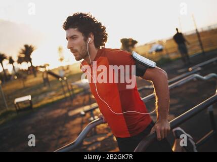 Portrait of a athletic young man listening to music on earphones exercising on an outdoor fitness park in the morning - young man doing dips outdoors Stock Photo