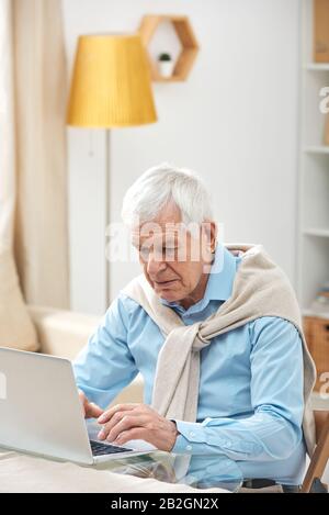 Serious busy senior man with sweated wrapped around shoulders using laptop while surfing net at home Stock Photo