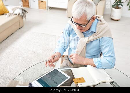 Content white-haired senior man sitting at glassy table and using tablet while analyzing own finances Stock Photo