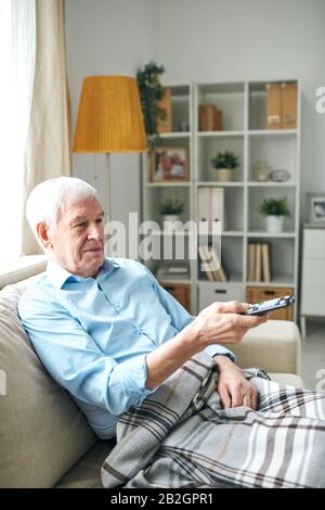 Content relaxed elderly man with blanket on legs sitting on sofa and using remote control while watching tv alone at home Stock Photo