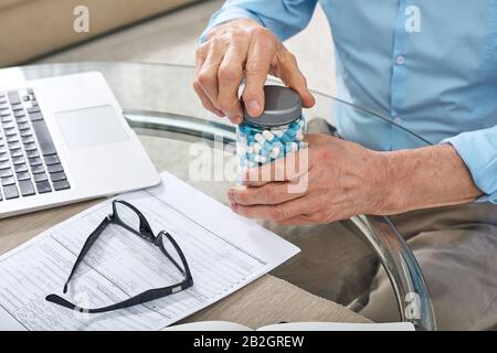Close-up of unrecognizable senior man sitting at table with laptop and opening pill jar while preparing to take medication Stock Photo