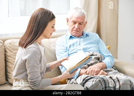 Smiling young female social worker sitting on sofa and reading book to elderly man under blanket Stock Photo