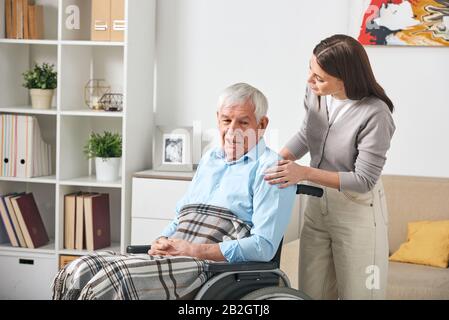 Careful young nurse talking to elderly man in wheelchair while visiting him at home Stock Photo
