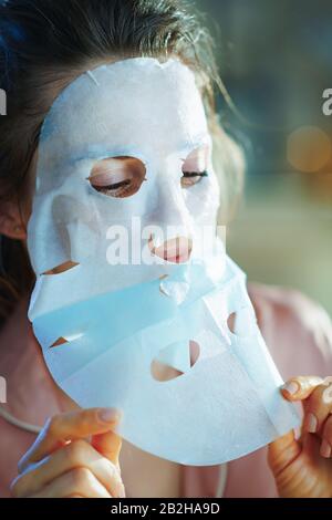 young female in pajamas in the modern living room in sunny winter day applying sheet facial mask and removing protecting layer. Stock Photo