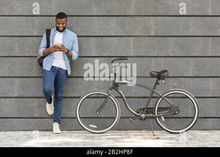 Young Black Man Using Smartphone, Standing Next To His Bike Outdoors Stock Photo