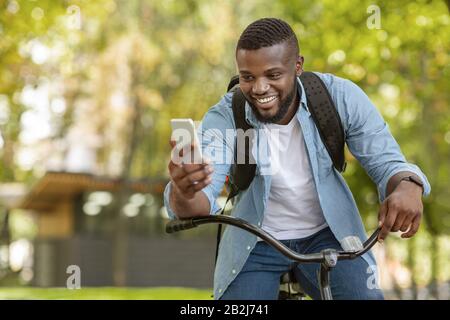 Happy Black Man Riding Bicycle Outdoors And Using Smartphone Stock Photo