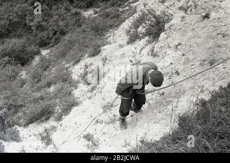 1950s, historical, young man absailing down a rocky cliff, England, UK. The technique involves using your body and the rope to make a controlled descent, using your feet and legs as support and balance as you lower yourself down. In this regard, he physical and mental skills needed to abseil, has seen become a popular 'adventure sport' or 'outdoor activity'. Stock Photo