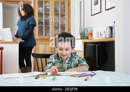 Boy (5-6) colouring in colouring book mother using laptop in background Stock Photo