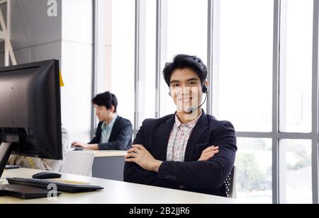 Smiling confident Asian male call center operator enjoy his work at office. Concept for customer support service, lifestyle at office, employee. Stock Photo