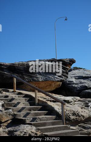 A single lone street lamp above and concrete steps below, the rocks of the ocean shelf and a wooden handrail leading to Maroubra Ocean Pool Stock Photo