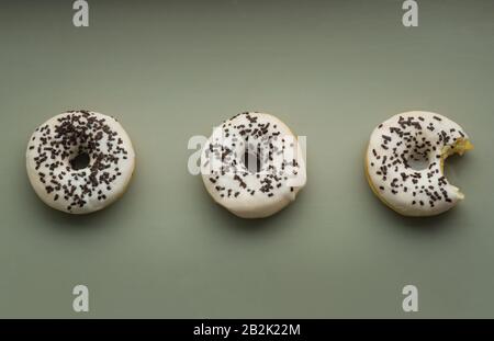 Three donuts with white glaze and chocolate sprinkles on a grreen or gray background, one of them bitten Stock Photo