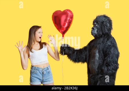 Young man dressed in gorilla costume gifting heart shaped balloon to surprised woman Stock Photo