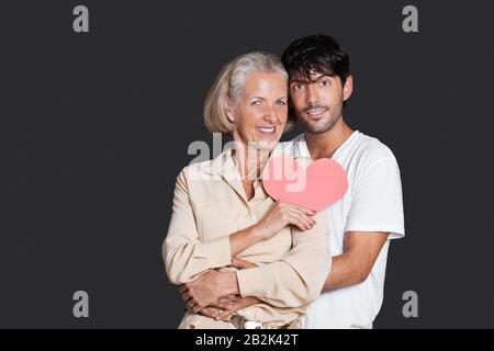 Young man embracing senior woman holding red paper heart against black background Stock Photo