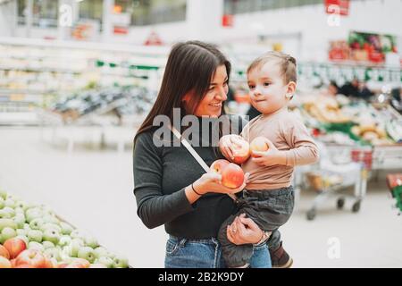 Young woman mother with cute baby boy toddler child on hands buys fresh apples in supermarket Stock Photo