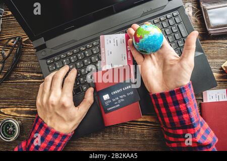 Preparing for an exciting journey. Man books flights by typing on a laptop and paying with a credit card on a wooden background. The view from the top Stock Photo