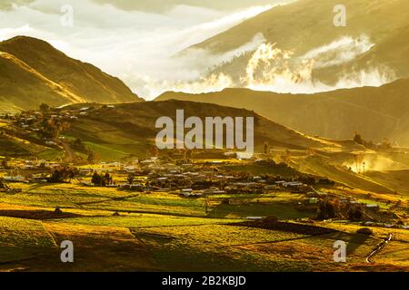 A rural village nestled in a mountain valley during golden hour, with scattered houses and farm buildings spread across rolling green hills. Terraced agricultural fields are visible in the Stock Photo